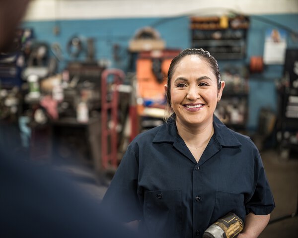 Frau in dunkelblauem Hemd steht lächelnd in einer Werkstatt  | © FOTOGRAFIA INC.